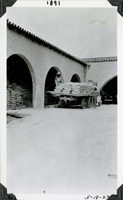 This is an historic black and white photograph from the Scotty's Castle Historic Photograph Collection, Death Valley National Park of flatbed truck loaded with bags of cement parked in large court yard. One man standing on back of truck, another man standing to side of truck. Building with open archways, cement bags inside. Inscriptions in black ink along upper and lower border.
