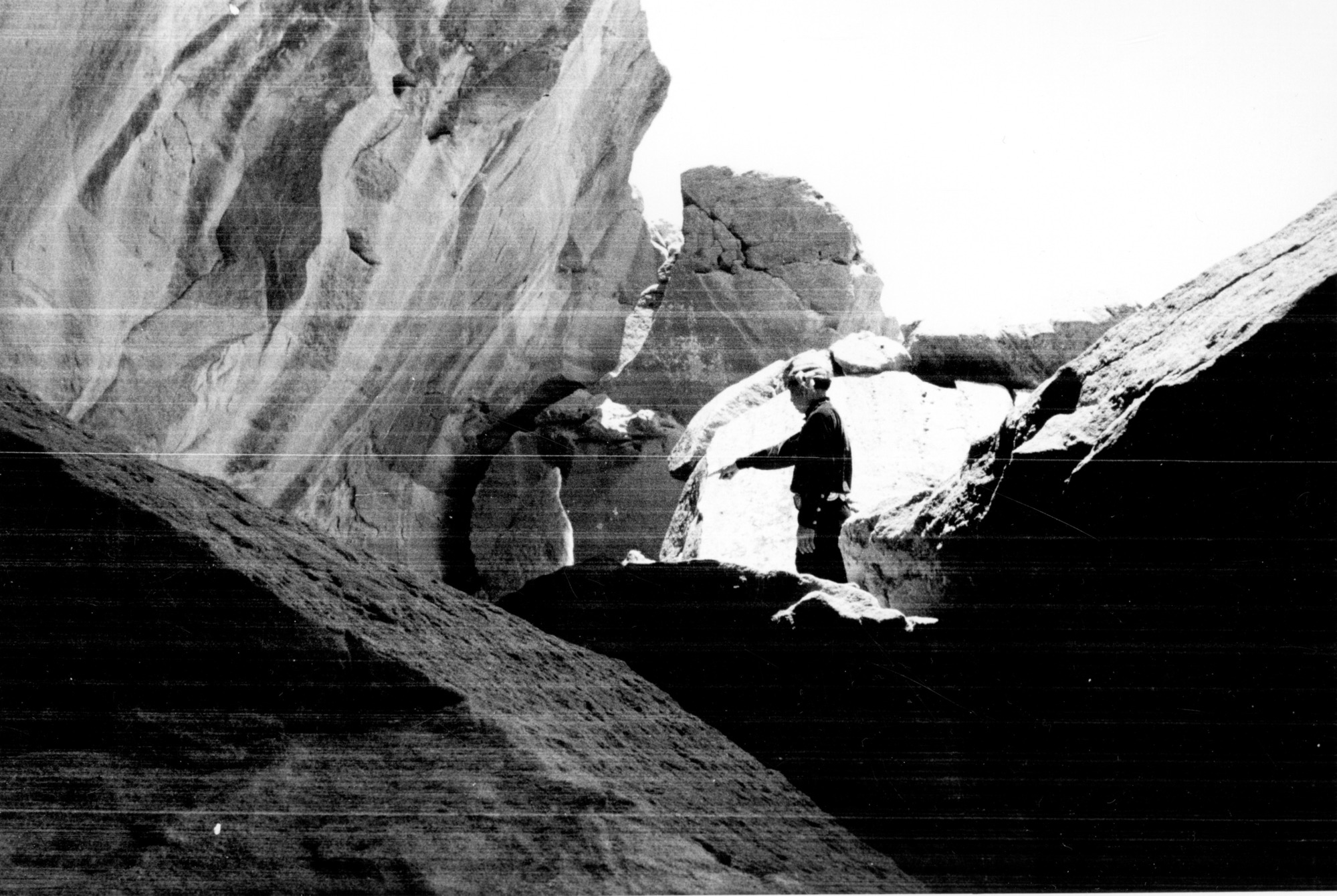 Black and white photograph inside of a narrow canyon. Porter Timeche is in the background pointing to a canyon wall, showing a petroglyph that is not visible. 