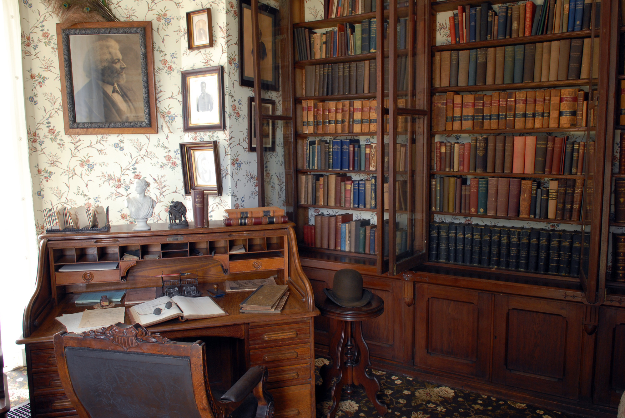 Two large bookshelves line a wall of a medium sized room. The shelves are packed with hundreds of books. A small wood desk sits in front of the book shelf. A photo of an older Frederick Douglass hangs on the wall. 
