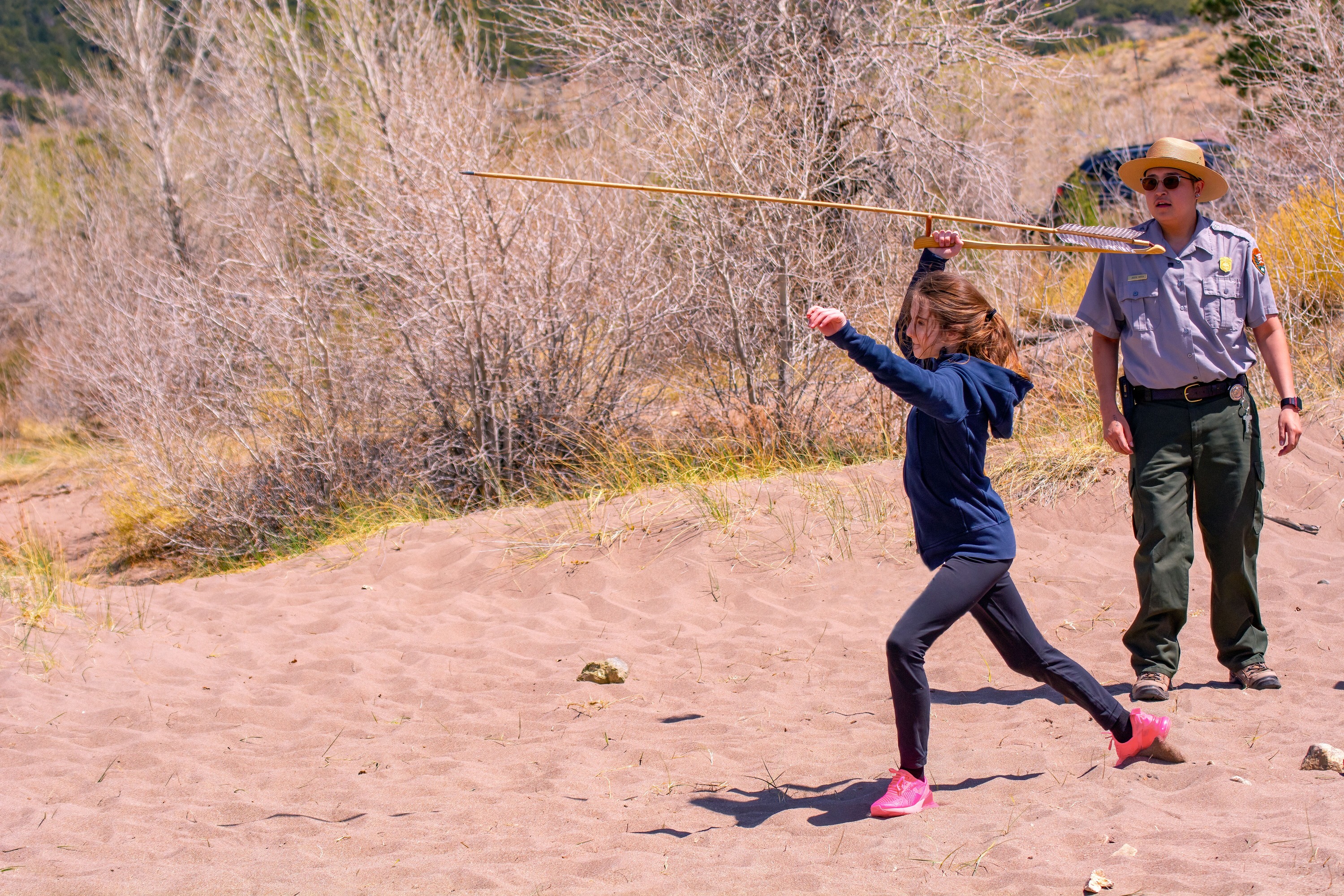 A student from Sangre de Cristo School launches a long dart with an atlatl during a presentation by Park Ranger Jaiden Garcia. They are both on sand with trees on the background.