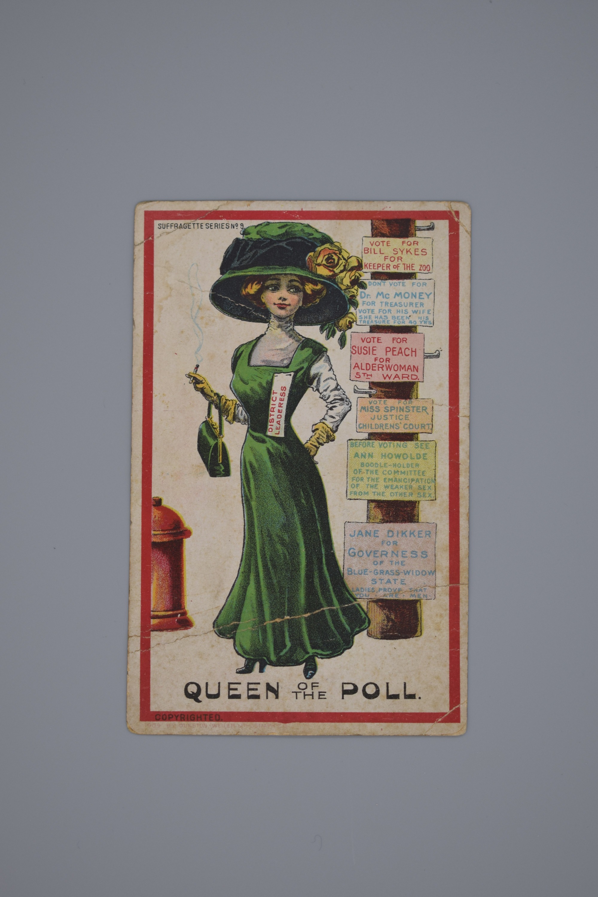 Postcard titled "Queen of the Poll" with illustration of a woman wearing a long velvet green dress with high-necked grey blouse, a large matching hat with black ribbon and yellow flowers, and yellow long gloves holding a green handbag and smoking a cigarette. Pinned to her dress above her chest on her left is a piece of paper which reads in red "District Leaderess" On her right is a red fire hydrant. On her right is a telephone pole with six signs attached. The signs text promote men and women candidates for different political offices including "Governess" and "Boodle-Holder of the Committee for the Emancipation of the Weaker Sex from the Other Sex."