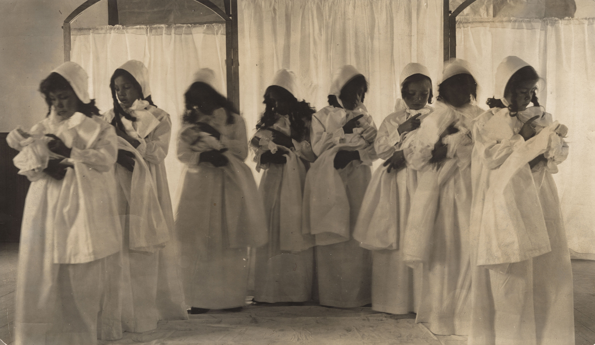 Black and white photograph of line of students dressed in white and holding dolls