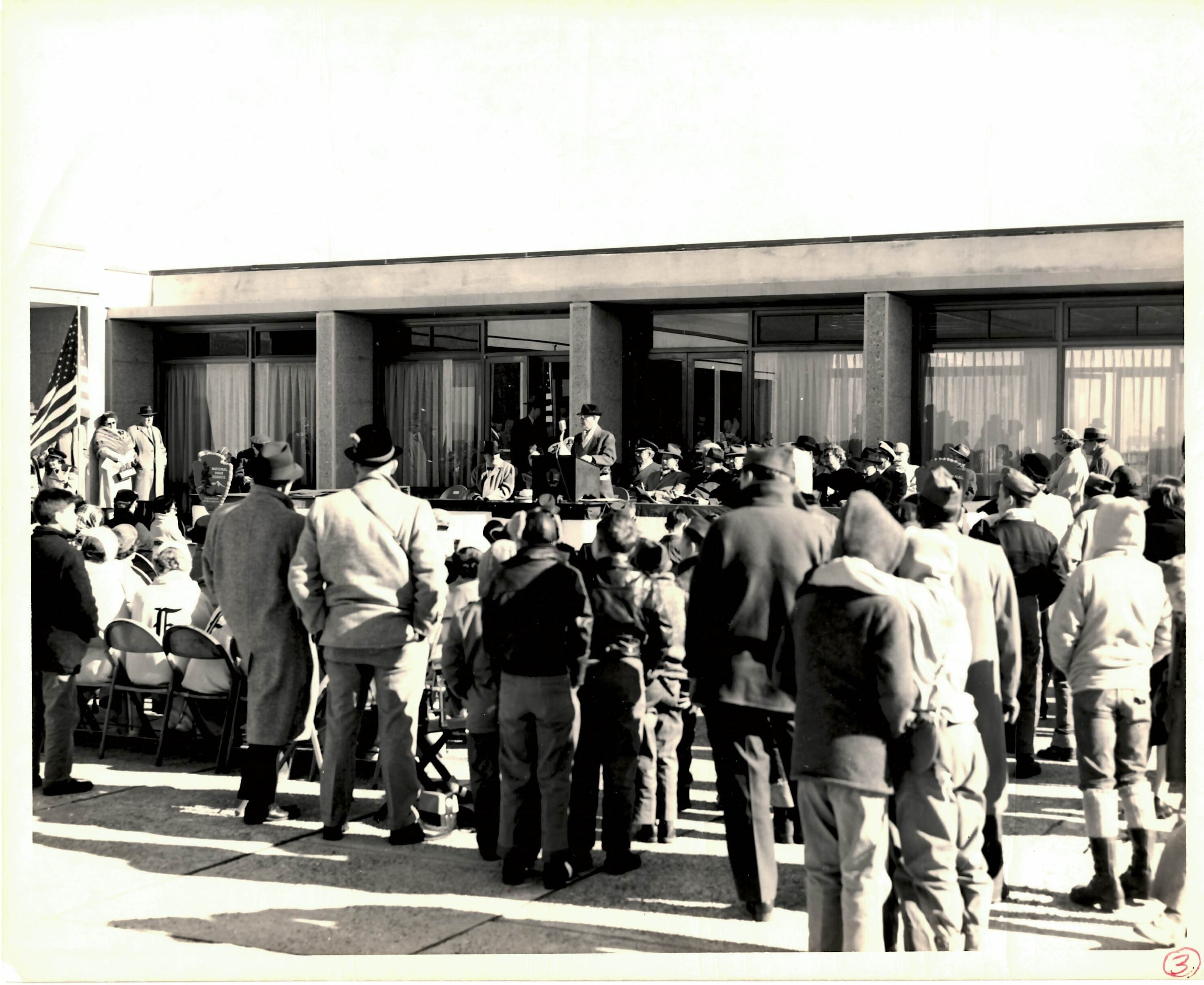 Black and white photo of crowd of people standing and sitting while listening to a man who speaks at a podium. Visitor center is in background. American Flag seen at left.