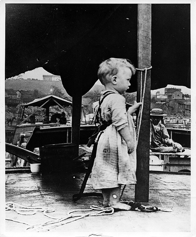 Black and white historical photo of a child secured to the canal boat using rope tied around her waist.