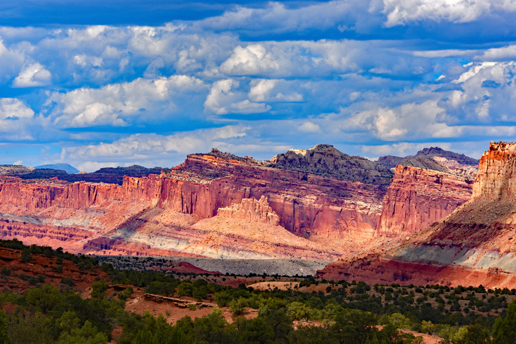 View of the Castle rock formation near an entrance to Capitol Reef NP. Formation includes striations with lignite and sandstone, red rock, and many stark stone features in front of blue and white clouds.