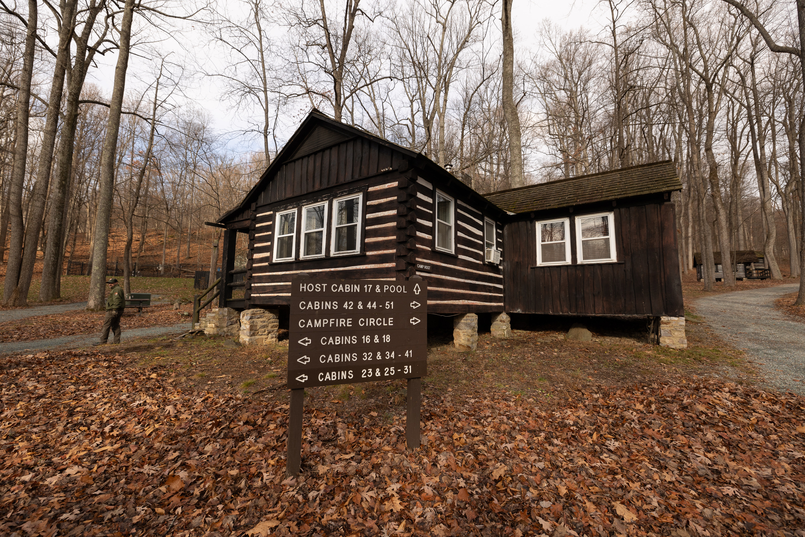 A directional sign reads, "Host cabin 17 & pool" and has an arrow pointing forward. "Cabins 42 & 44-51" has an arrow pointing to the right. "Campfire circle" has an arrow pointing to the right. "Cabins 16 & 18" has an arrow pointing to the left. "Cabin 32 & 34-41" has an arrow pointing to the left. "Cabins 23 & 25-31" has an arrowing pointing to the left. The directional sign is positioned in front of a large cabin. 