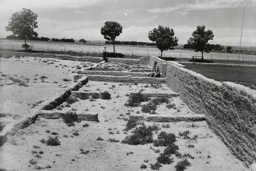 Two men stand or sit among low adobe walls placed to define the perimeter of Bent's Fort following archeological work in the 1950s. These walls were not historic and not always accurate in their display of room alignment. A row of trees lines a driveway that circled the site of the fort. 