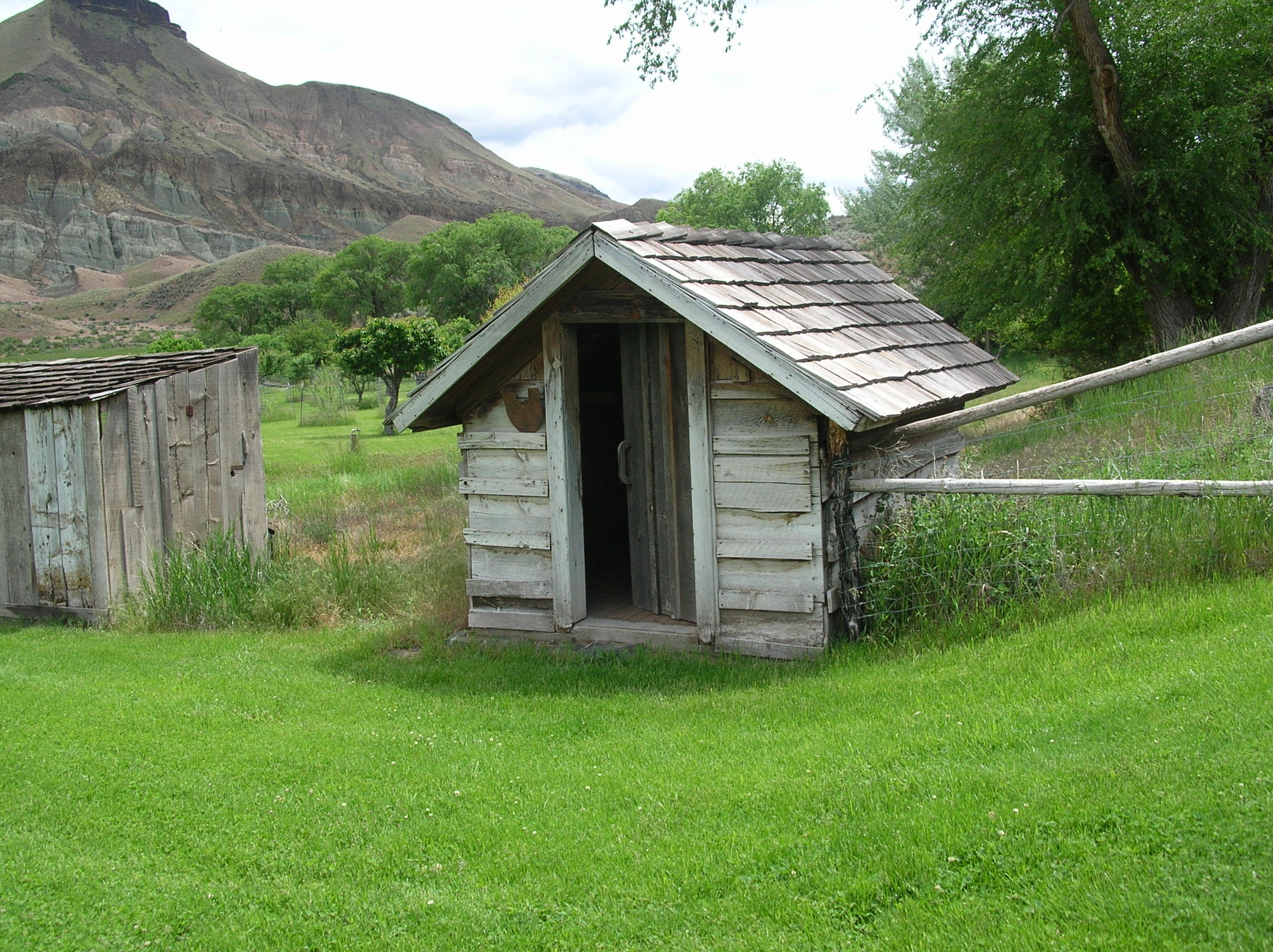 A small shed sits at the corner of fencing on bright green turf with trees and rock formations beyond. 