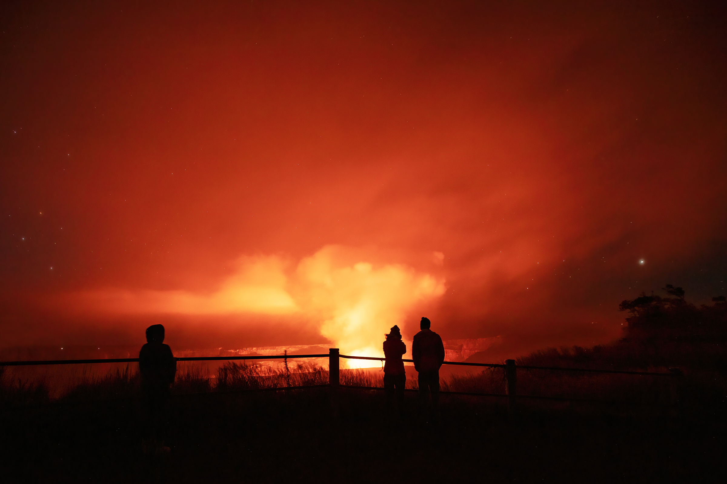 Silhouettes of human figures looking at a orange glowing volcanic plume and crater at night