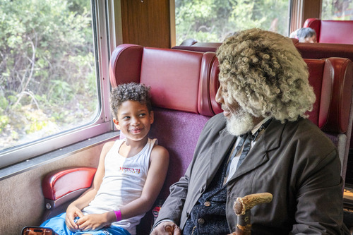 A young boy smiles up at an older man dressed in a 19th century suit and gray wig