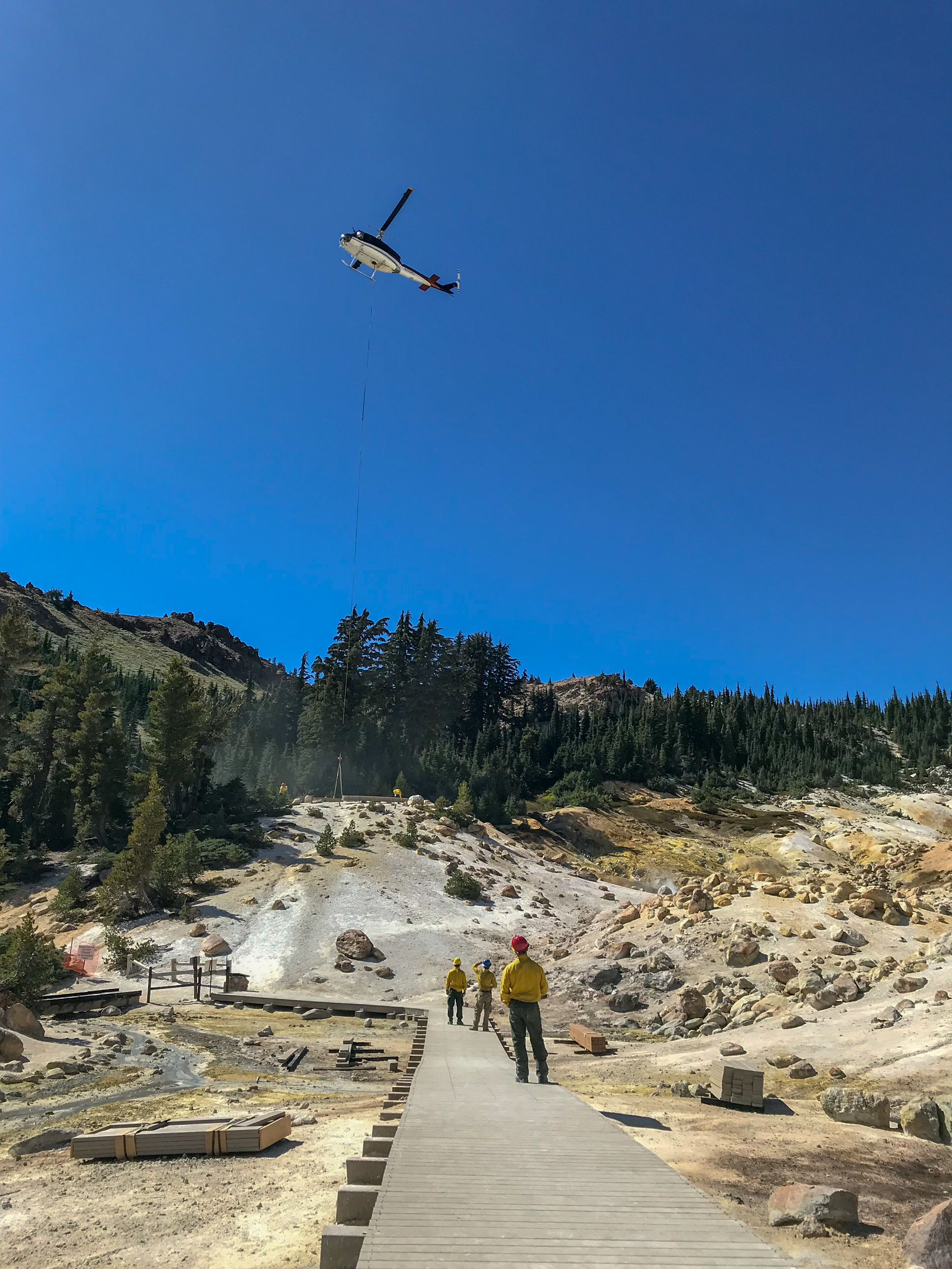 Three people in yellow shirts and hardhats stand on partially completed boardwalk while a helicopter hovers overhead in a blue sky