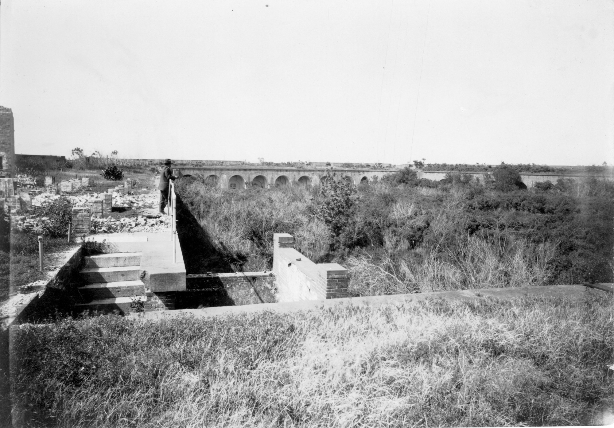 Look at the parade ground of a brick fort that is overgrown with trees. 
