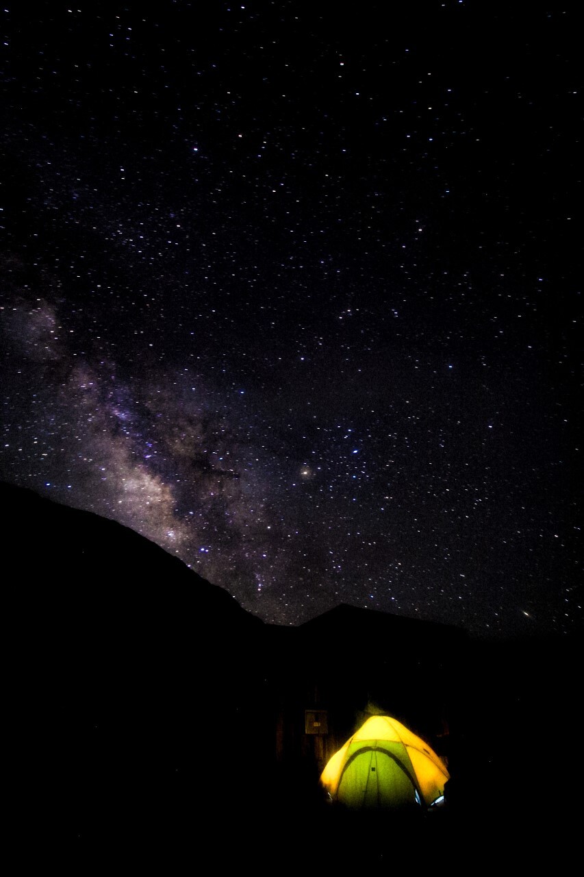 A yellow tent glows from the inside below a starry night sky.