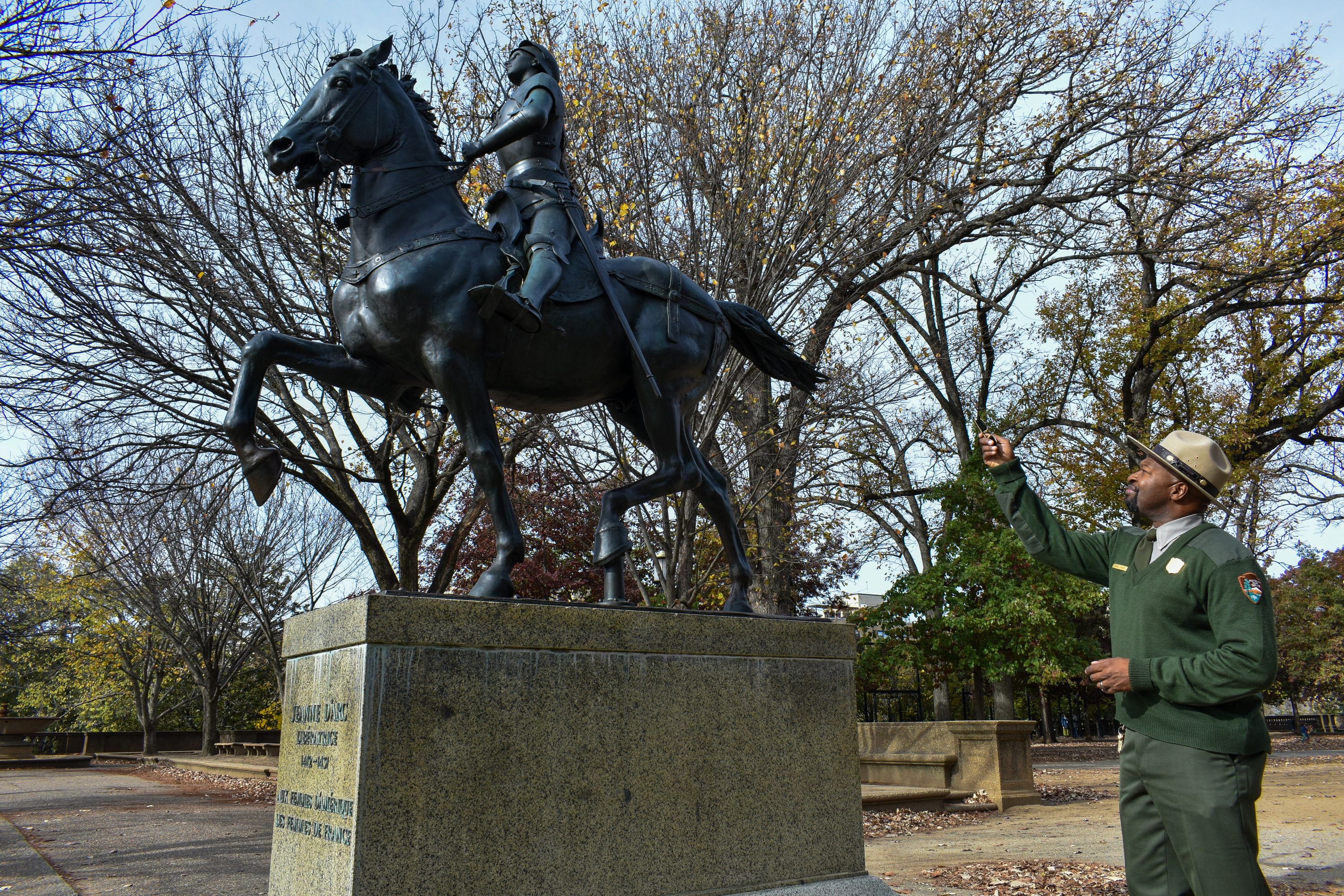 A man in National Park Service uniform stands beside and points at a large statue of Joan of Arc sitting on top of a horse.