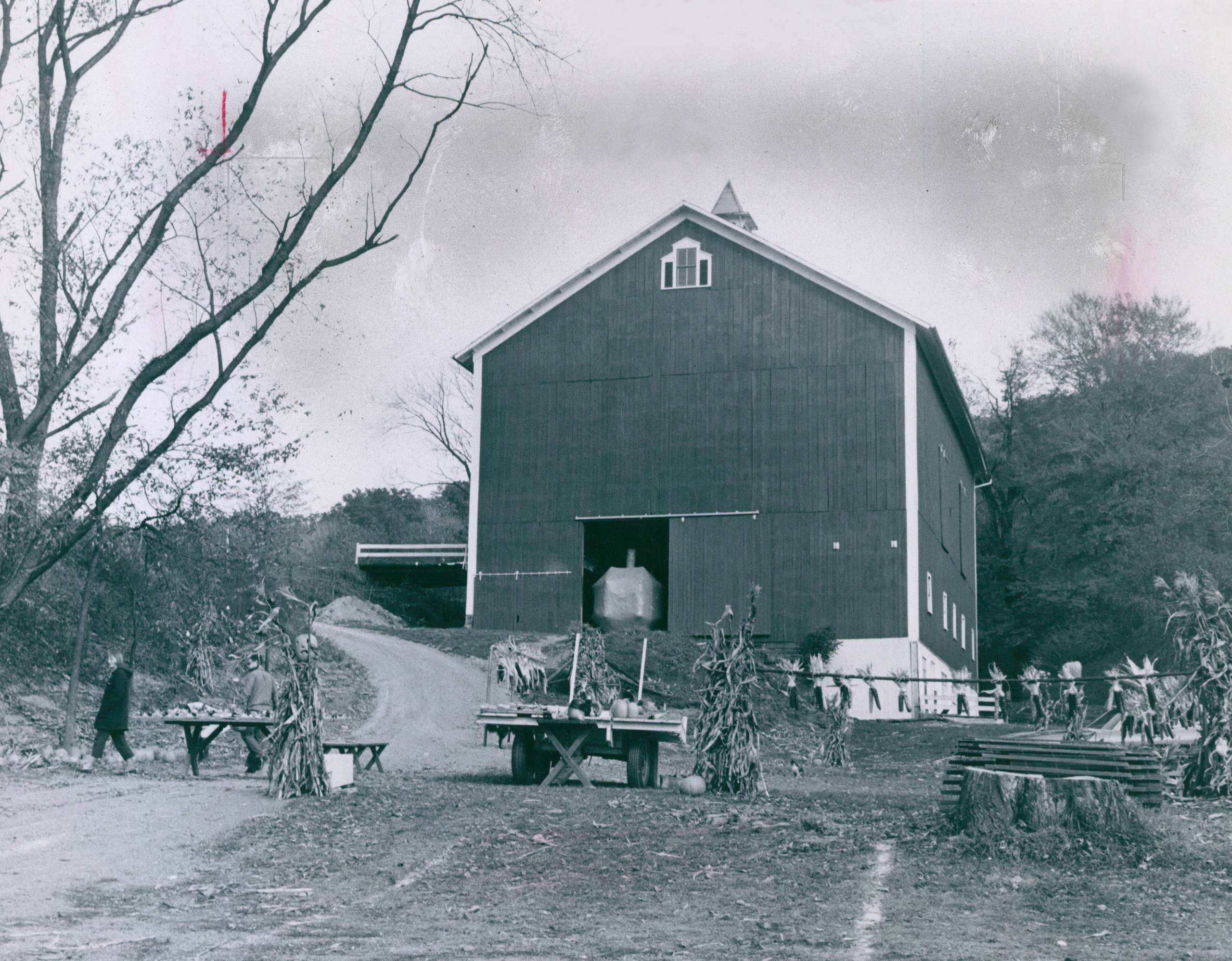 Black and white photo of a large, multi-story bank barn with a peaked roof, a farm stand set up in front near the dirt driveway.