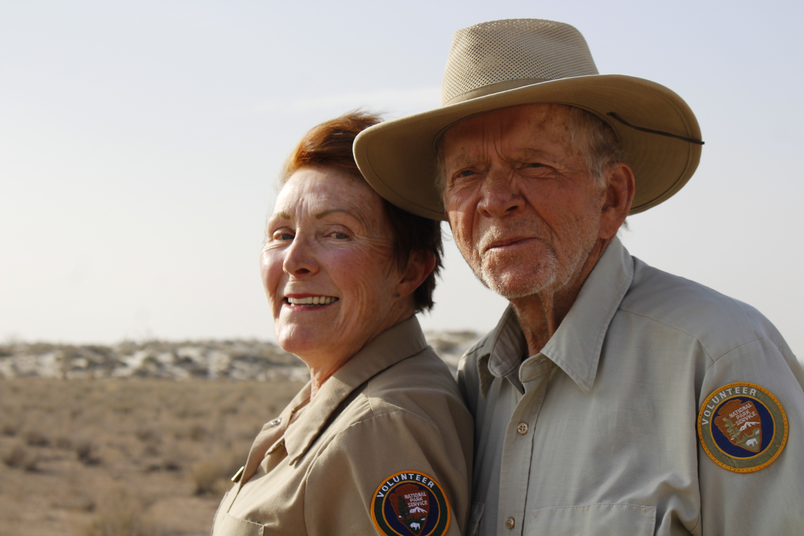 A man and a woman stand side to side dressed in the tan NPS volunteer uniform. The man is wearing a hat and grinning towards the camera. The woman looks to the camera and smiles. The desert unfolds in the background meeting white sand dunes covered in plants under a pale blue sky.