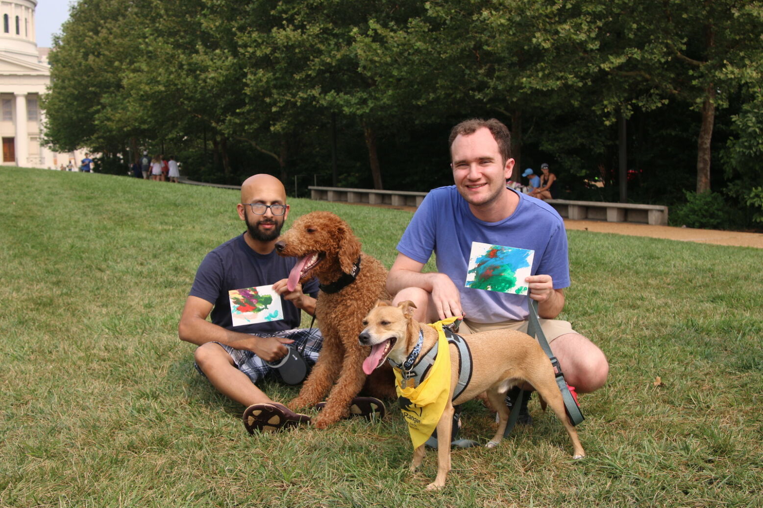 Two humans hold small canvases of artwork while their dogs stand in front of them with tongues wagging.