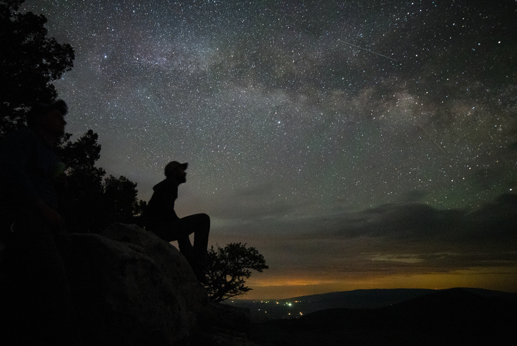 A silhouette of a person wearing a cap sitting and looking up at a star-filled sky.