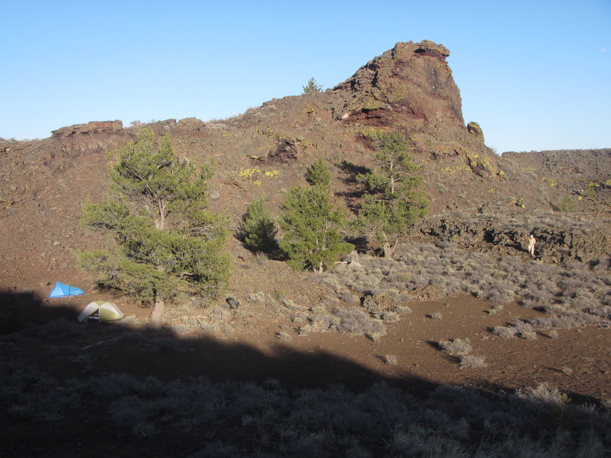 two tents and a person next to the wall of an open crater lined with some sagebrush and four pine trees