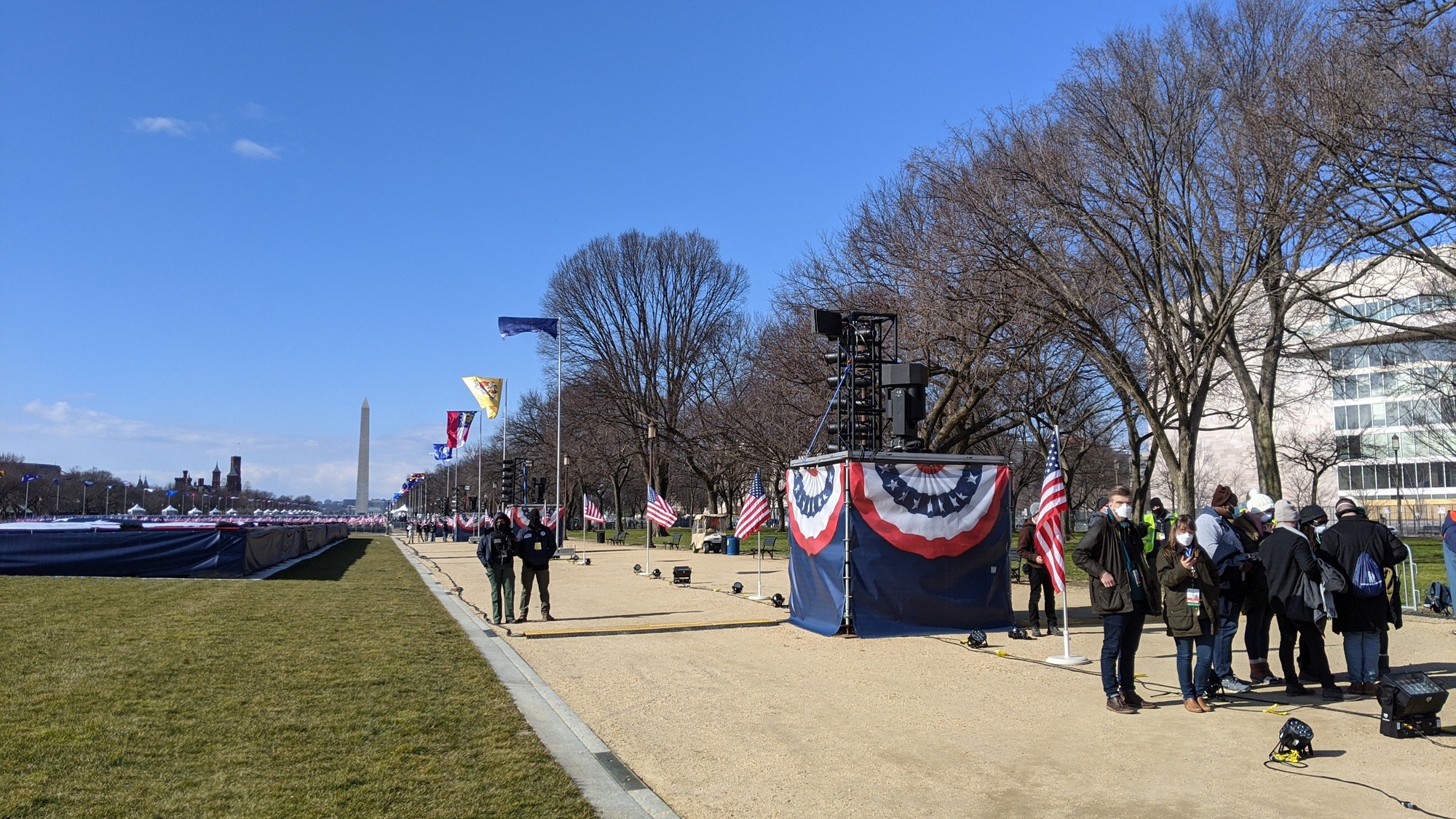 A wide pathway leading towards the Capitol is lined with spaced lighting stands, event staff, and state flags on flagpoles. A rectangular blue structure holding up a horizontal U.S. flag is on the grass lawn next to the pathway. The Washington Monument is in the distance.