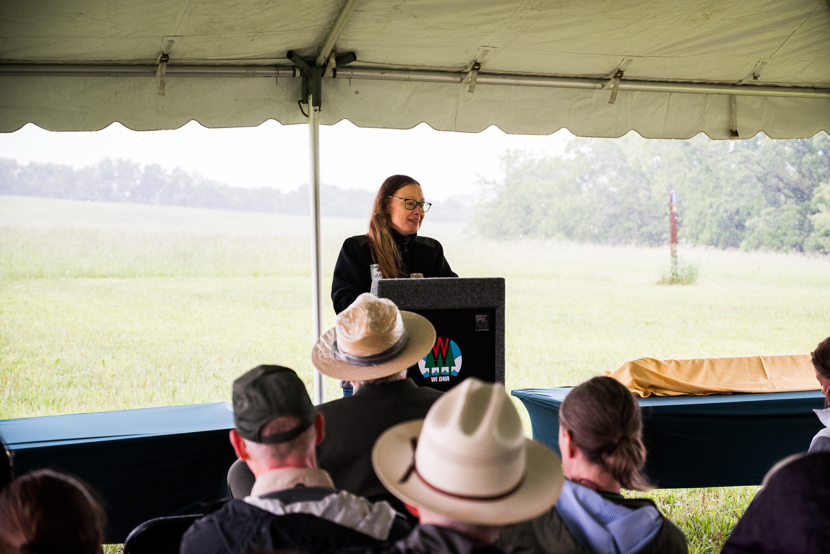A photograph of a person wearing a black sweater speaking at a podium under a tent. The background shows an outdoor, rainy scene.
