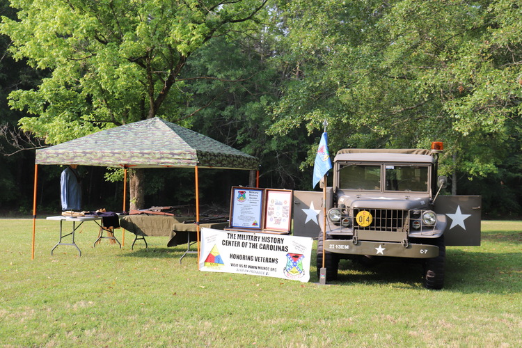 An olive drab Military truck sits next to a camouflage tent with tables holding guns from WWII and has a banner setup in between them with information.