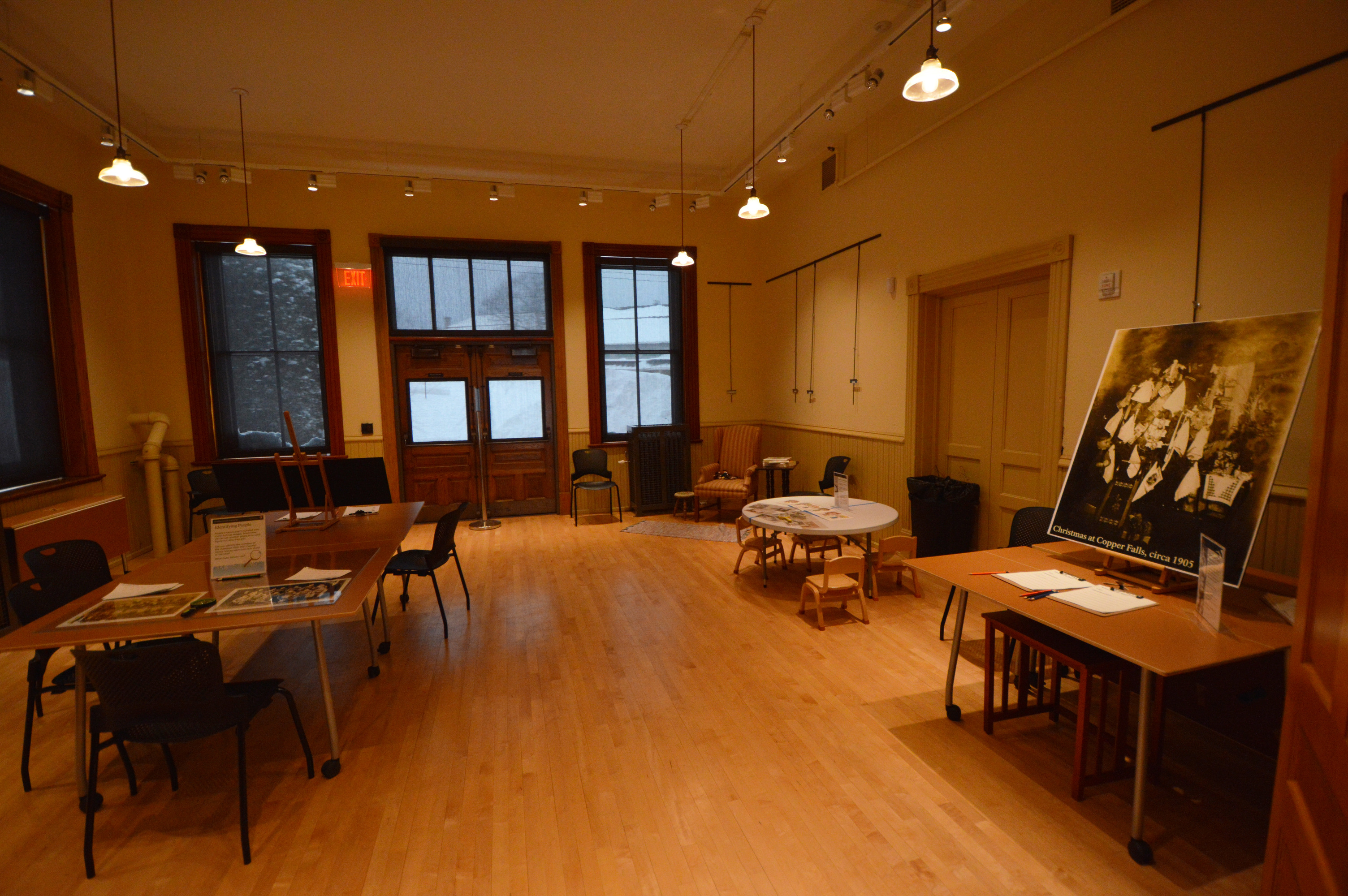 A wood floor room with several tables of various sizes spread throughout the room. plastic chairs are spread around tables. The tables have posters, clipboards or other materials for visitors to interact with. A tall back upholstered chair is in the back right corner. Two emergency double doors in the center of the back wall open to the back of the building. 
