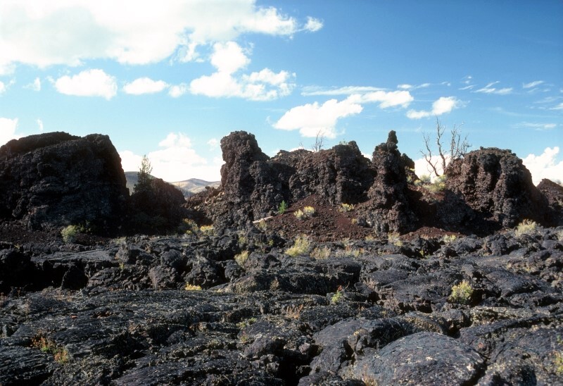 a bluish black lava flow with sparse vegetation in front of several large black and reddish rocky outcrops
