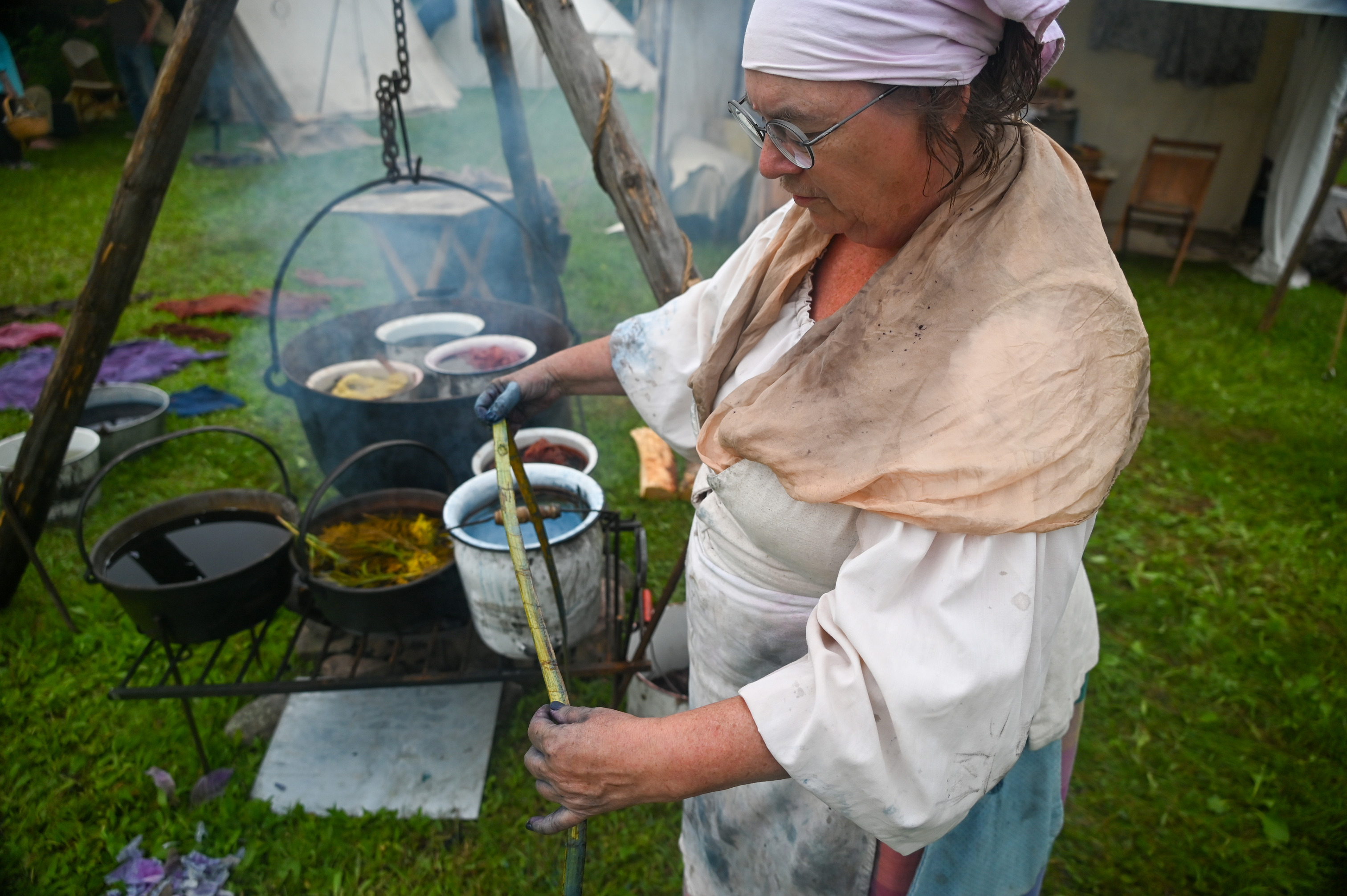 A women holds out a cedar strip displaying its new changing color. Behind pots heat over a fire in a rainbow of different colors.