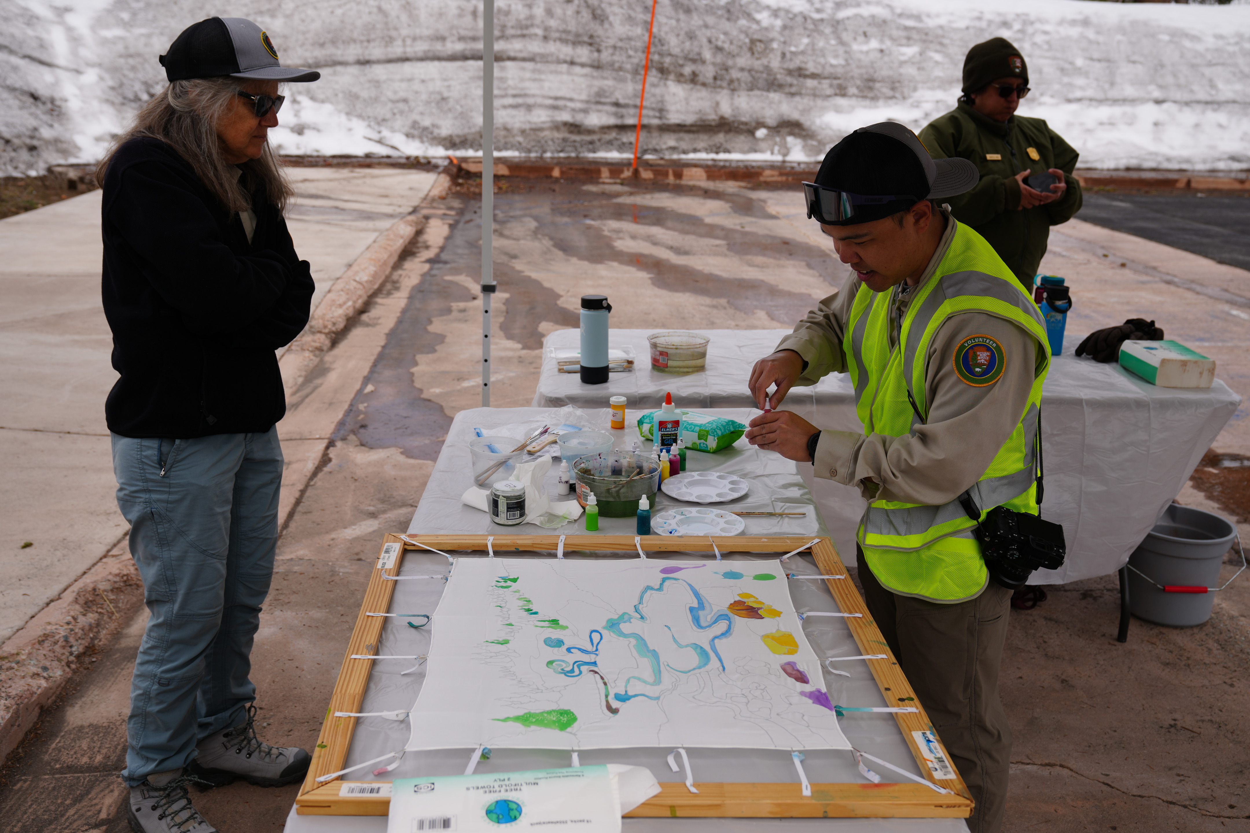 Volunteer unscrews red paint bottle over silk canvas to help paint.