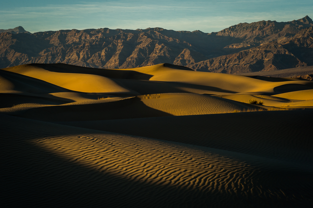 Golden sand dunes, with heavy shadows and wave patterns in the foreground and bare mountains in the background.