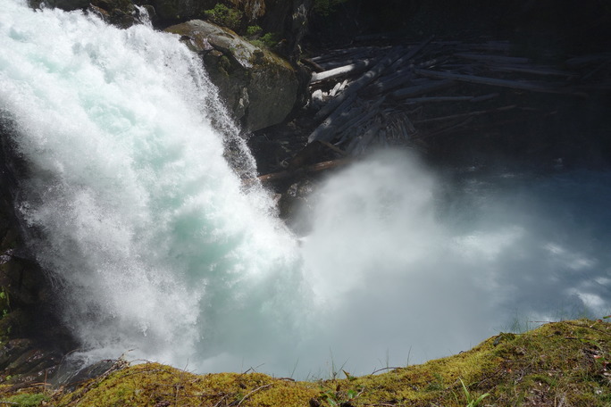 A torrent of blueish-white water roars over a cliff, sending up a large cloud of spray. 