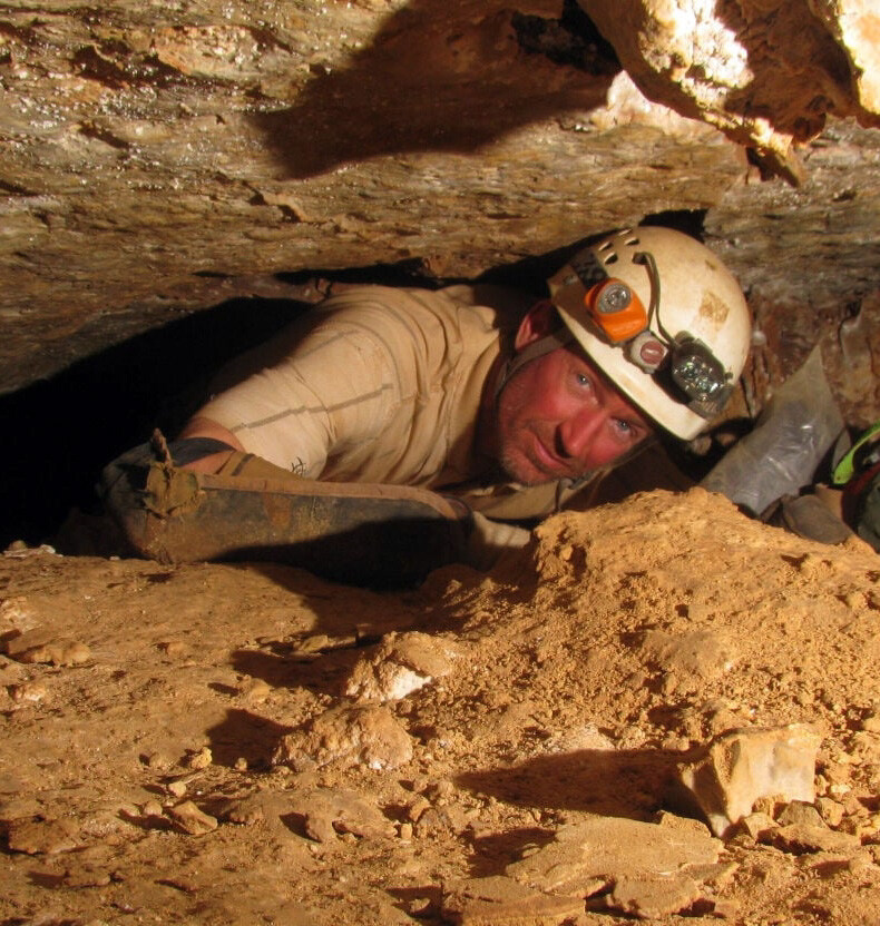 A man inside a small cave chamber, wearing a safety helmet with two lights.