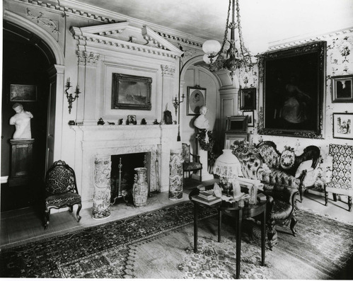 Black and white photograph of elaborately decorated 19th century parlor, featuring paintings, sculpture and chandelier.