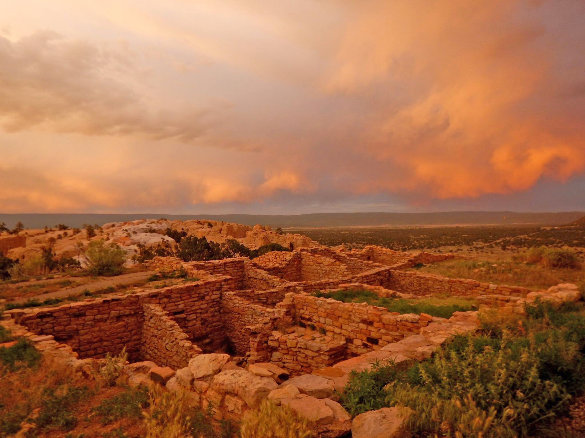 Threatening clouds are hit with the orange light of sunset with the square rooms of Atsinna pueblo in the foreground and the zuni mountains in the background. 