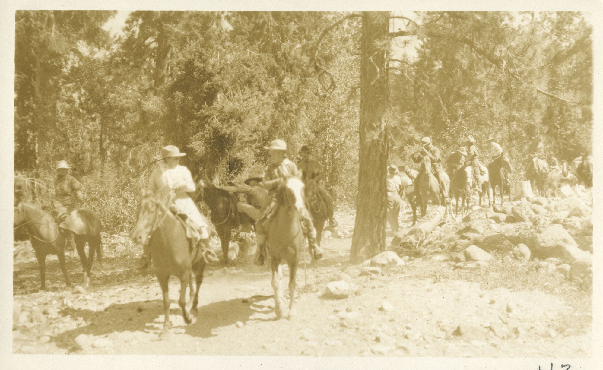 Horseback riders, led by Mary Roberts Rinehart and male riders, cross rocky meadow under Ponderosa pine.