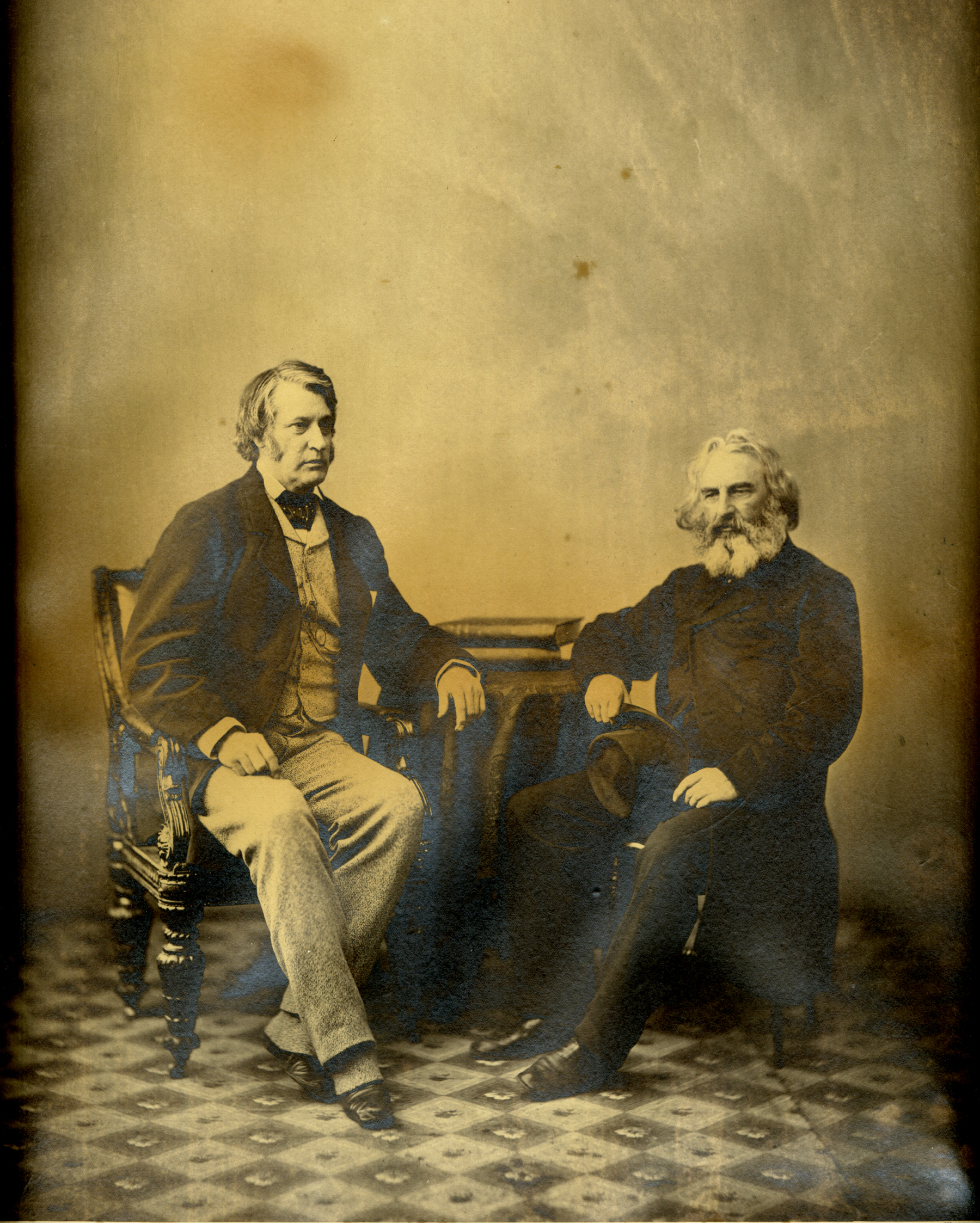 studio portrait of two men sitting next to small table, one very tall and sitting upright, one with long white beard