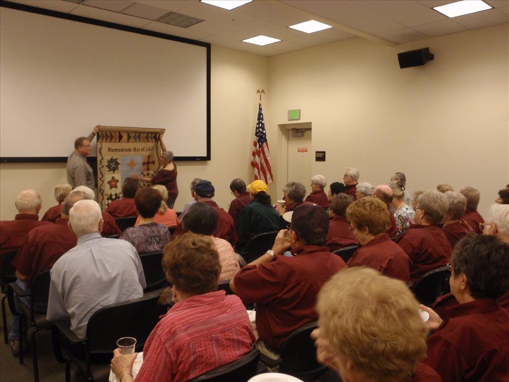 A group of volunteers sit in a room watching the unveiling of a quilt.