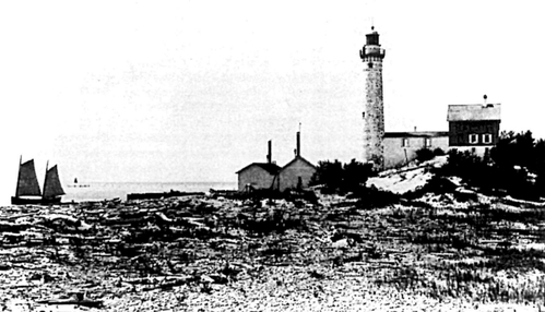 A grainy black and white photograph with sand and vegetation in the foreground. On the left, in the background, you can see a ship with two sales on water. On the right, are two small buildings with two stacks sticking straight up, and a lighthouse with a long structure attaching it to a small building.