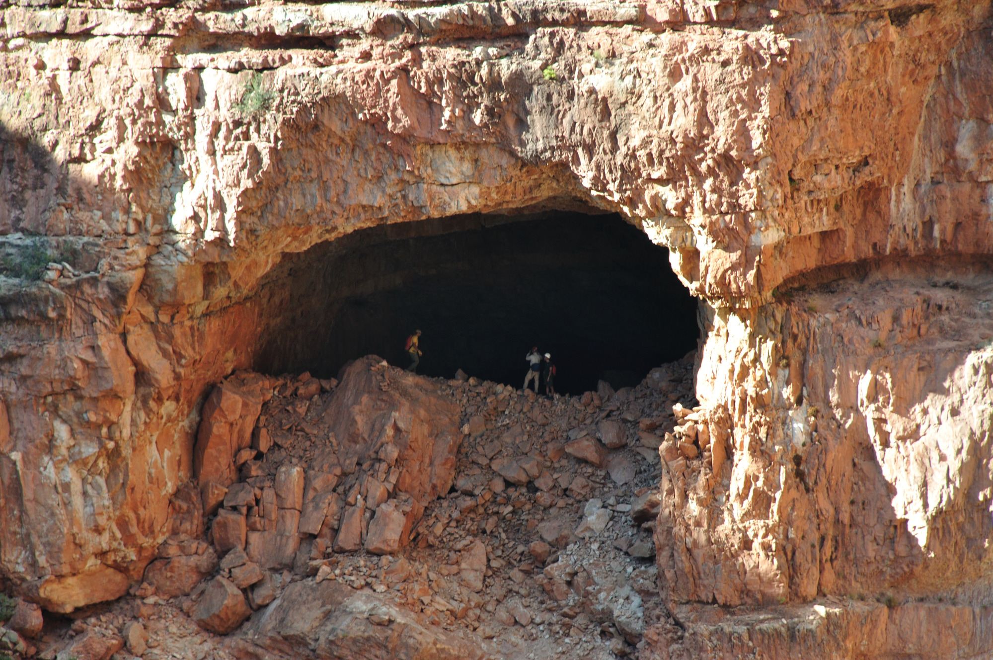 3 people stand in a large opening to a cave in the side of rocky cliff.