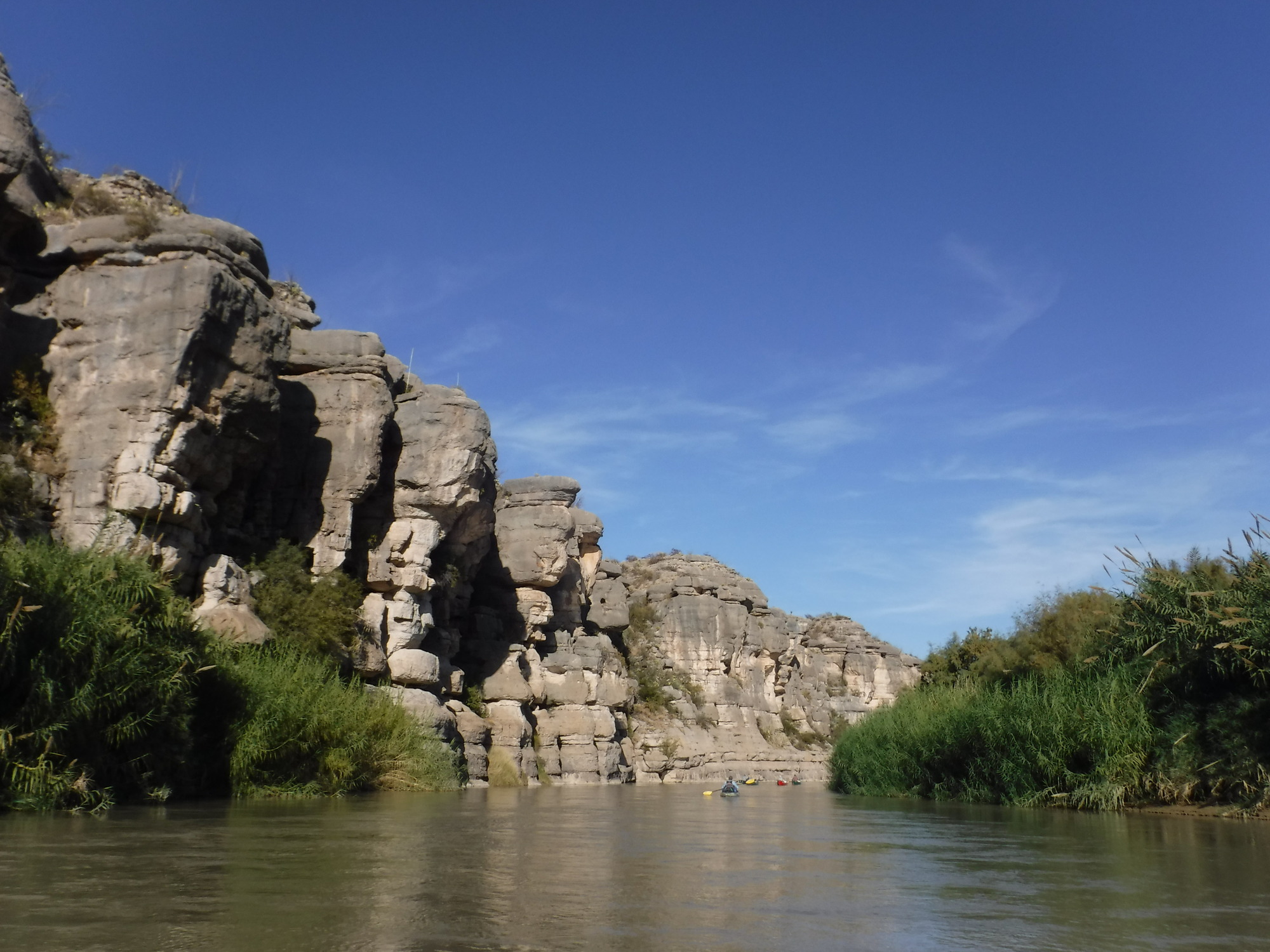 A view of the Rio Grande from the perspective of a paddler looking downstream. Two boats on the far horizon drift down a calm reach of water with giant cane growing on the right side and dramatic cliffs cut with vertical faults to the left. The sky is blue save for two or three wispy white clouds.