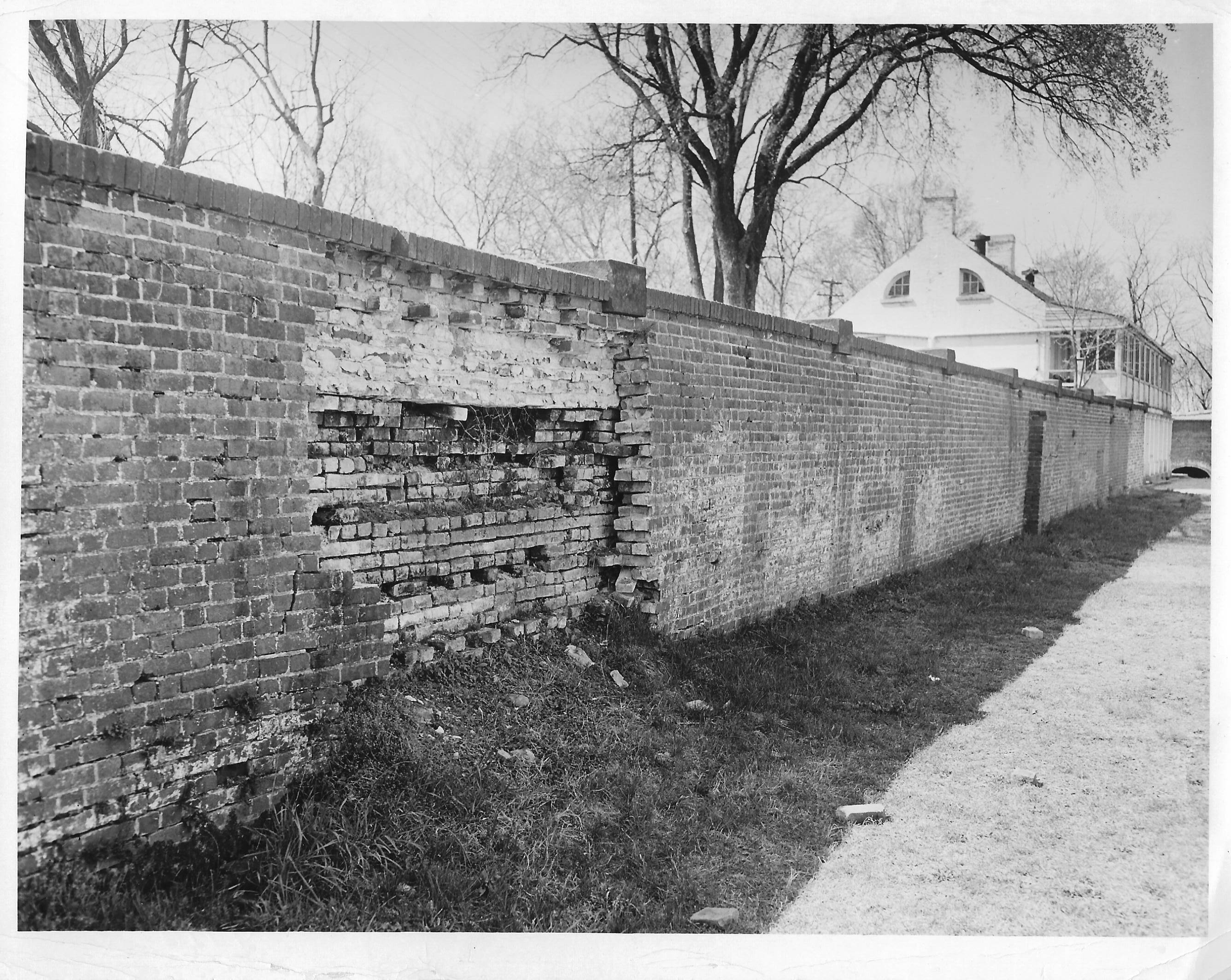 A section of the wall has been damaged, the outer layer of brick is missing, showing an inner layer of brick which is mostly intact.  A pile of dirt is below the damaged area.  The Enlisted Men's Barracks are at the end of the wall.