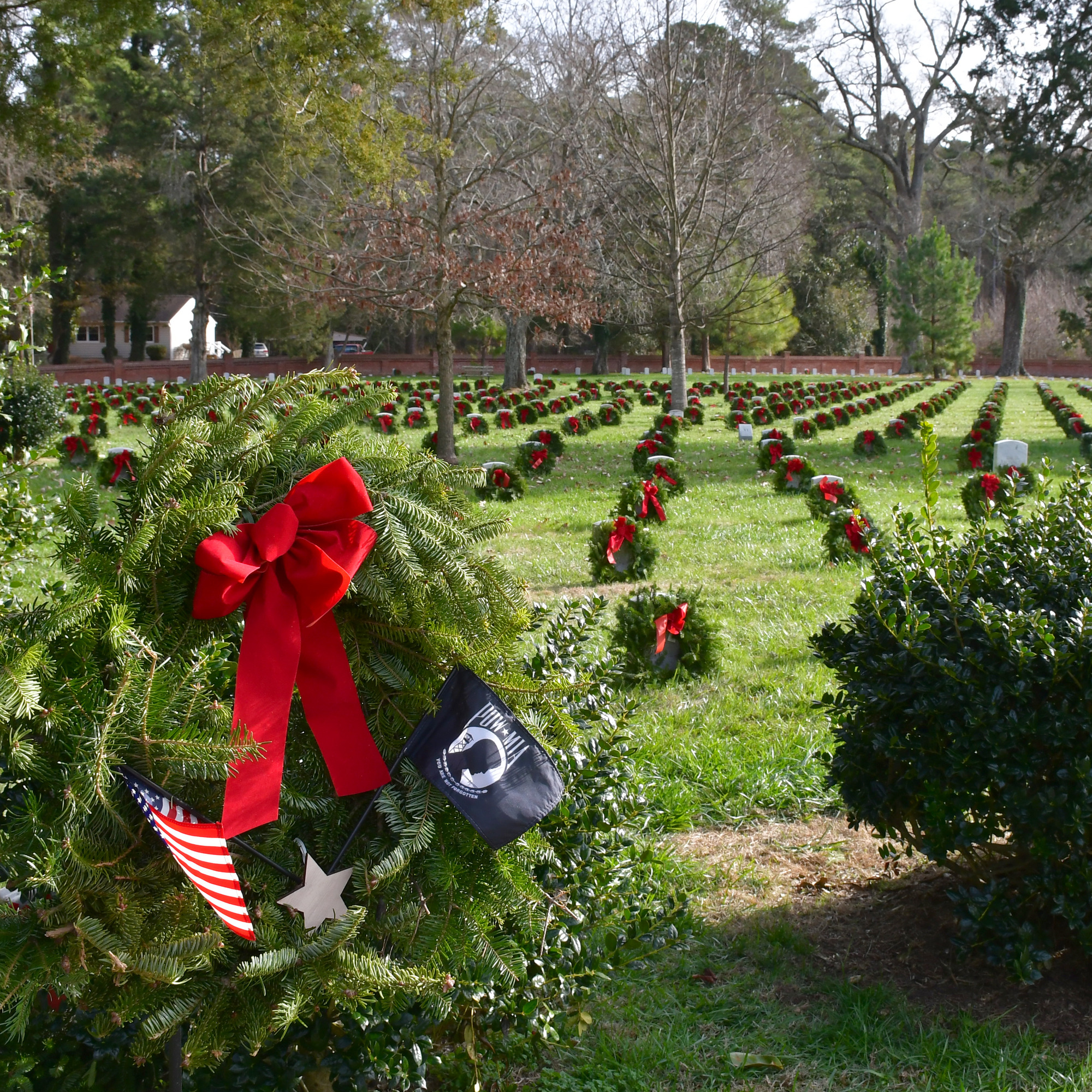 Close up of a green wreath with a red bow, U.S. flag, and POW/MIA flag; hundreds of headstones adorned by green wreaths with red bows fill the background