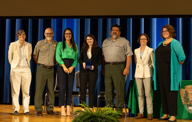Seven people in ranger uniforms and buisness attire pose for a photo on stage during an awards ceremony.