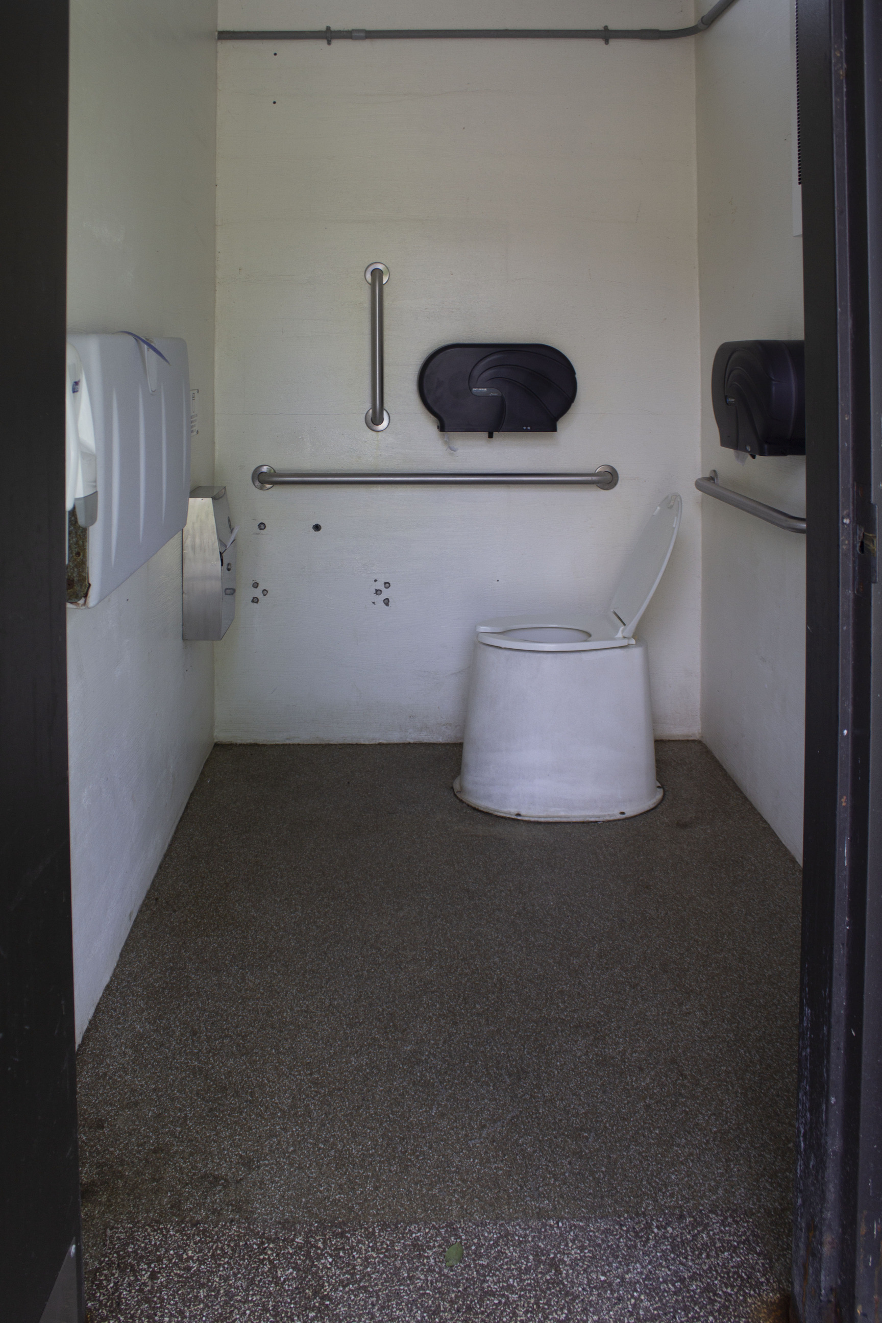 Interior of a vault toilet; two toilet paper dispensers are mounted above metal grab bars on two of the walls.