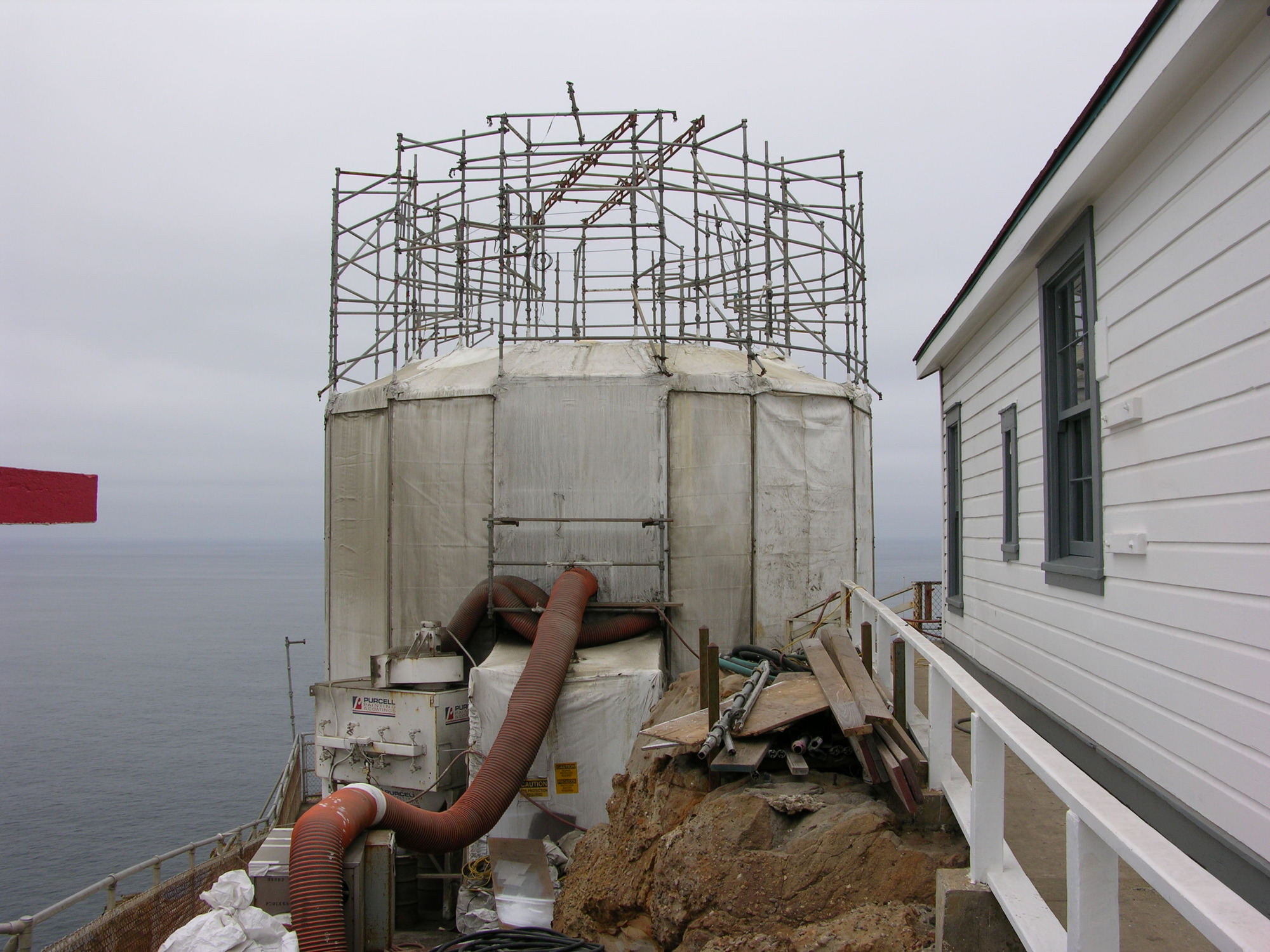 A cylindrical, white but dirty, nylon shroud and metal scaffolding surrounds sits on a rocky headland above the Pacific Ocean. Large flexible tubes connect a ventilation system to the "shroud" surrounding the tower. On the right is the side of a wooden building painted white with gray window frames.