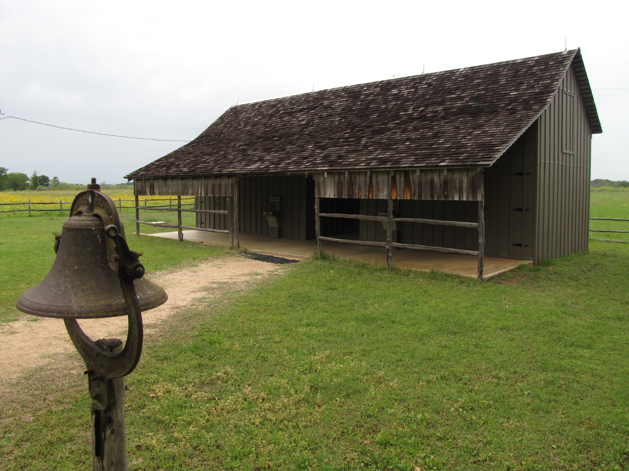 Wooden barn-looking building surrounded by green grass stands behind a rusted bell on a post.