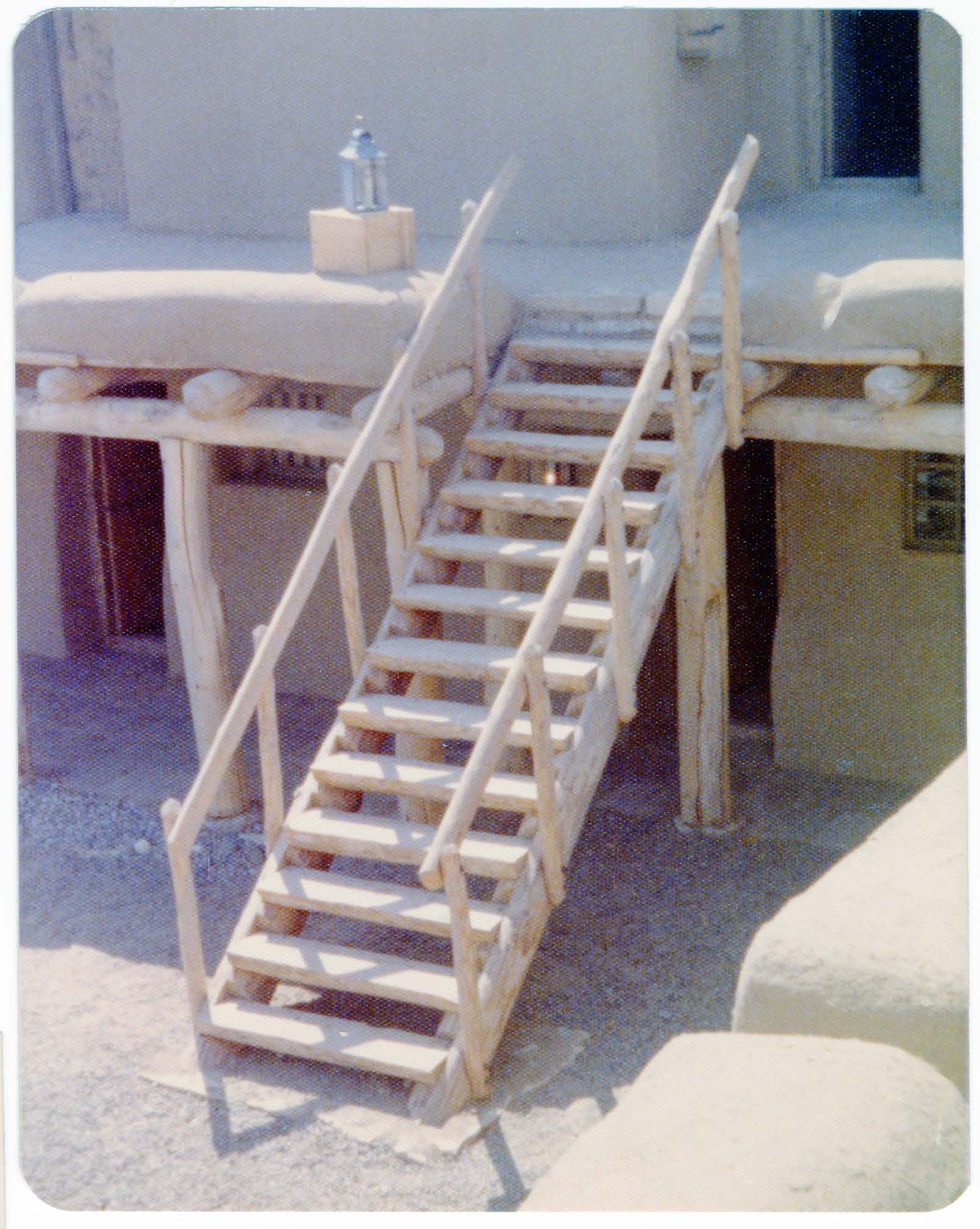 Taken from the location of the watchtower, the wooden staircase with handrails is seen leading from the plaza up to the west range of rooms. 