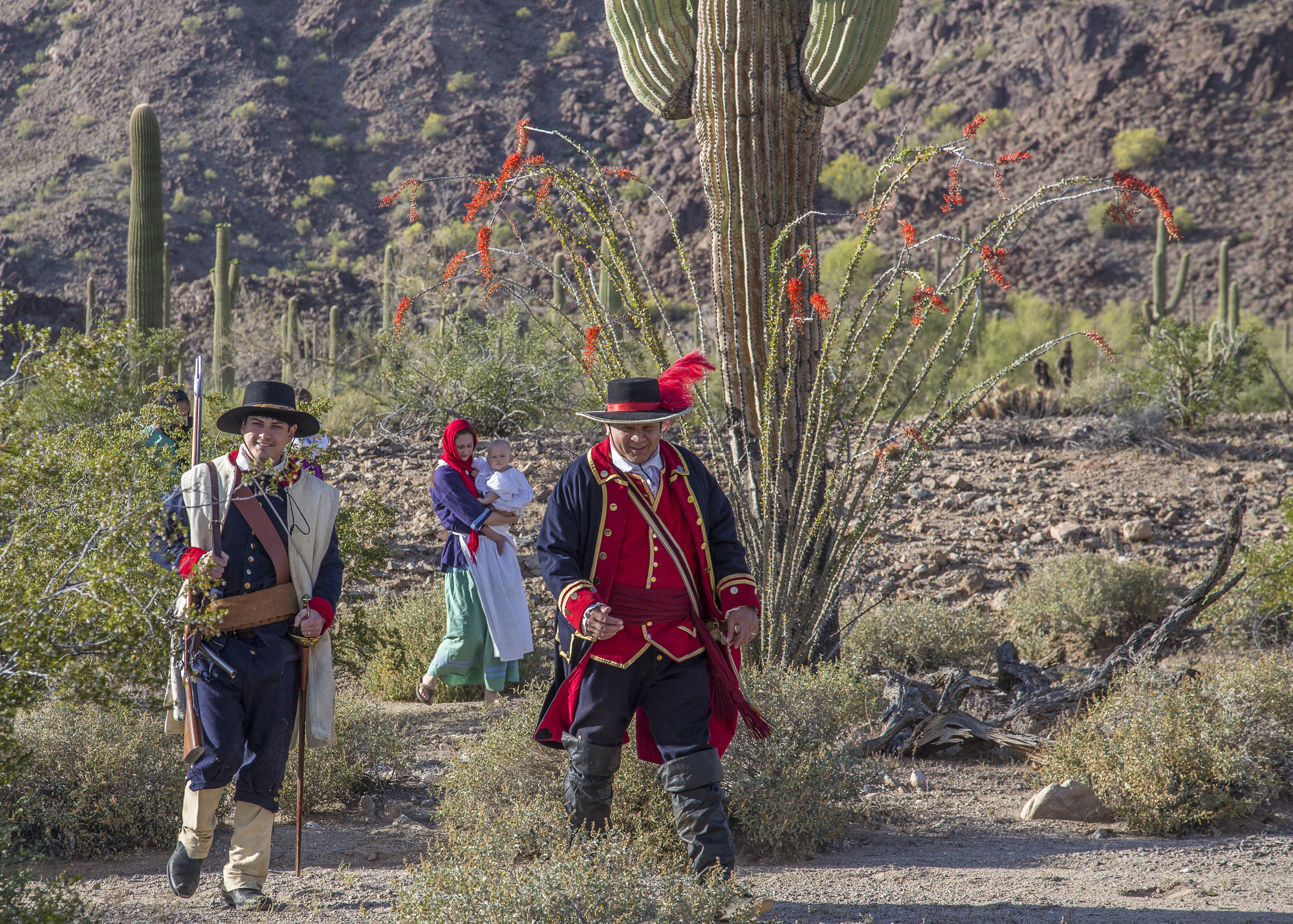 Two men wearing Spanish colonial military uniform and two women carrying children walk through the desert