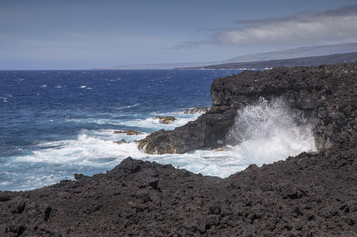 Waves crashing a long a sea cliff. The southern point of Hawaiʻi can be seen in a distance. 